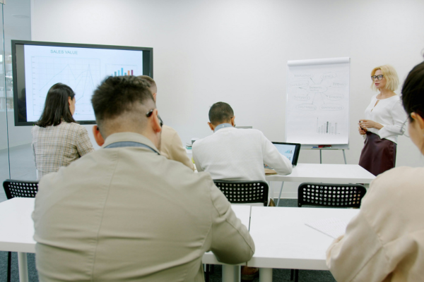 A small group of people sitting at long desks in an office environment with someone presenting at the front of the room